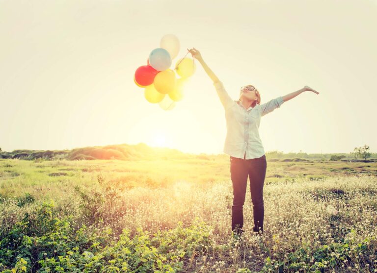 Frau mit Luftballons freudig die Arme hochstreckend auf einer Wiese in der Sonne