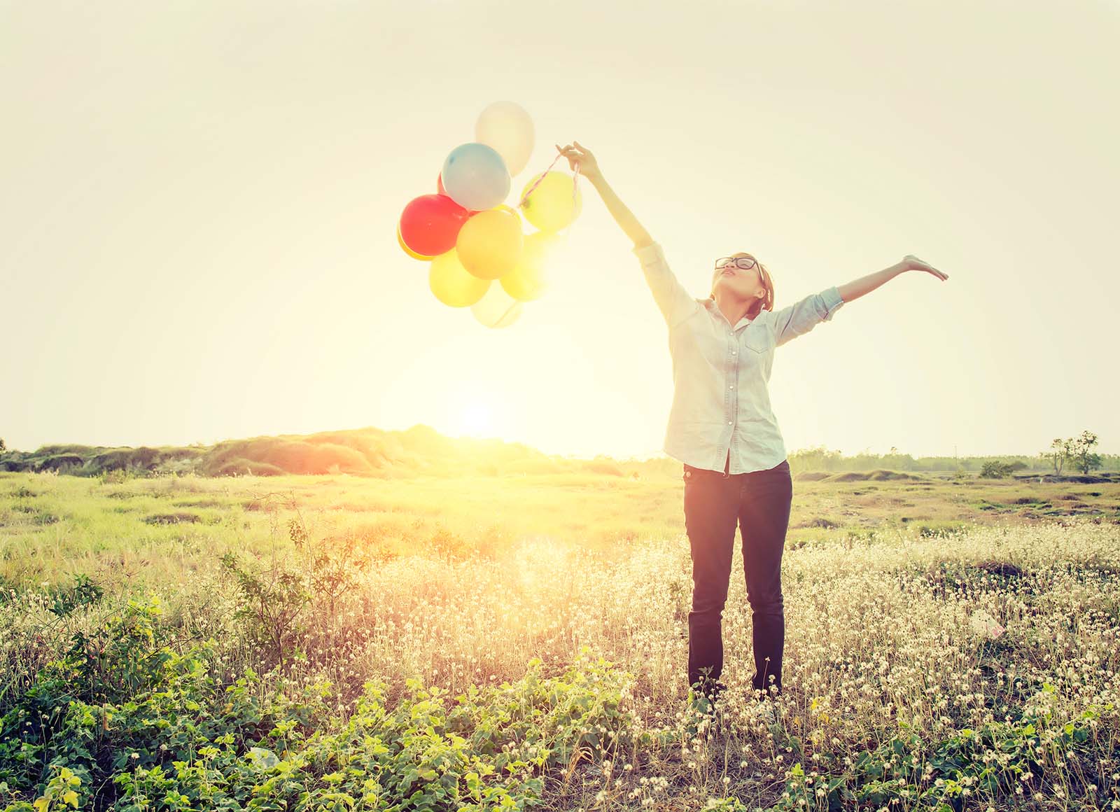 Frau mit Luftballons freudig die Arme hochstreckend auf einer Wiese in der Sonne